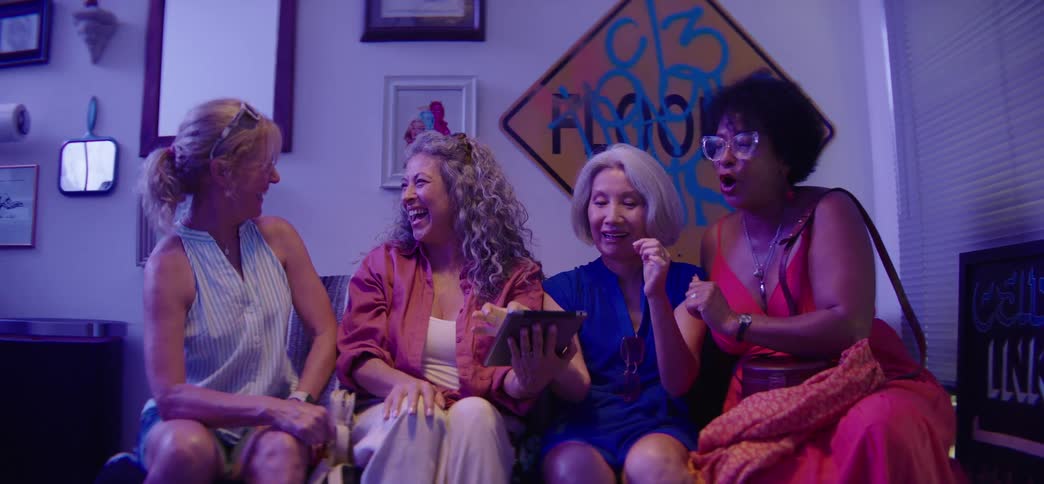 A diverse group of fun-loving senior women friends sit together on a couch, laughing and reacting with joy as they watch something on a tablet