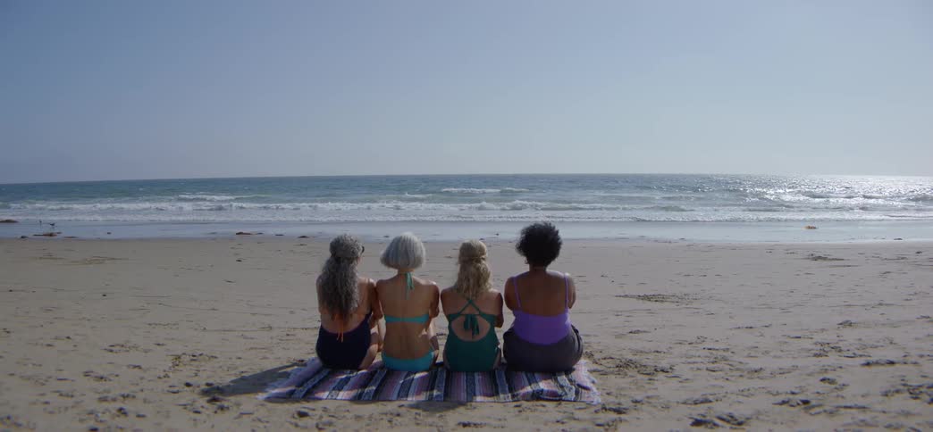 Four diverse senior women sit peacefully together on a beach blanket, enjoying the tranquil ocean view on a sunny day