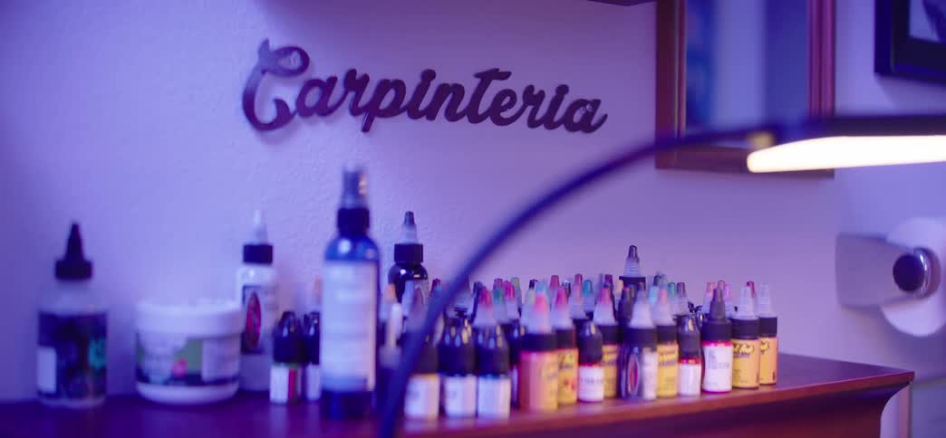 Colorful bottles of tattoo ink and supplies line a shelf in a Carpinteria studio under a cool purple light