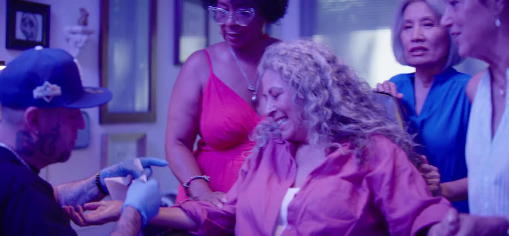 A group of diverse older women friends gather to support one of them getting her first tattoo at a tattoo parlor