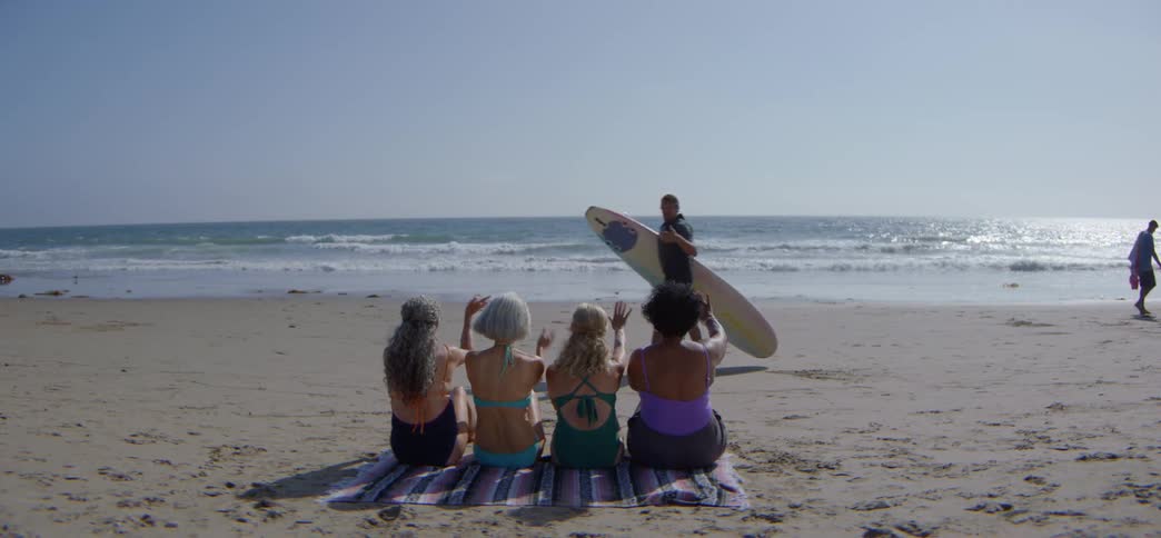 Four senior women in bikinis sit on a beach towel and wave at a young surfer walking by on a sunny day