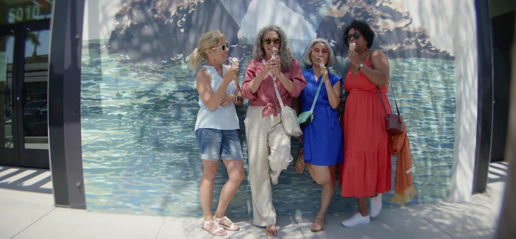 A diverse group of four happy senior women friends laugh together while eating ice cream cones against a beach mural on a sunny summer day