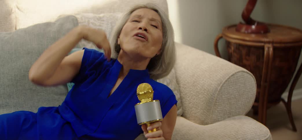 A dolly shot from a sleeping dog to an elderly Asian woman singing karaoke with a golden microphone while relaxing on her couch