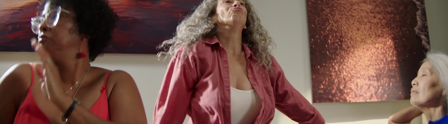 In slow motion, a joyful senior woman with long curly gray hair dances freely while her friends look on with unamused expressions