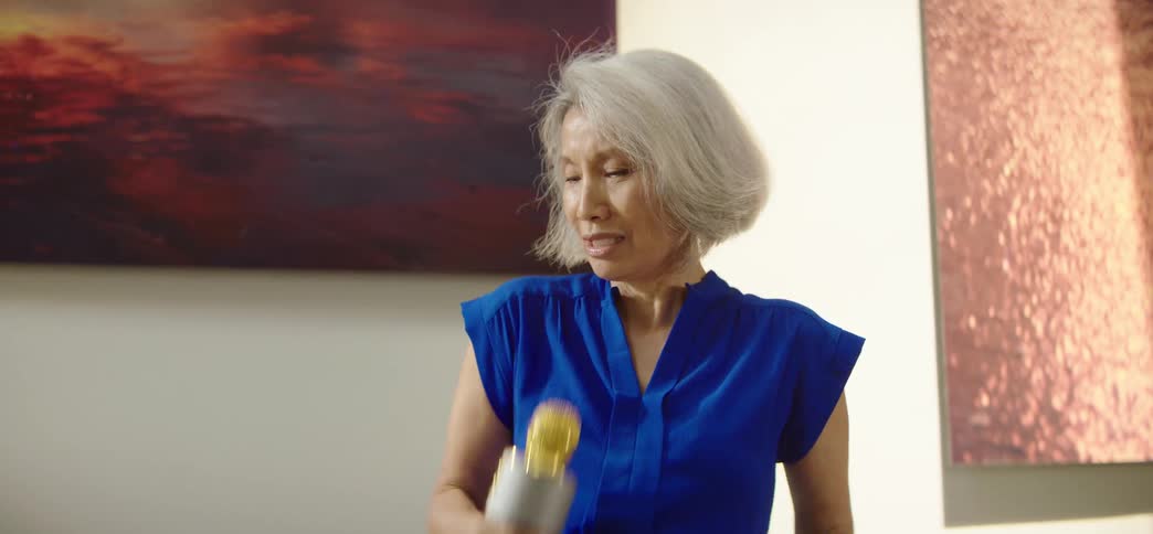 An energetic elderly Asian woman sings and dances with passion using a toy microphone in her living room