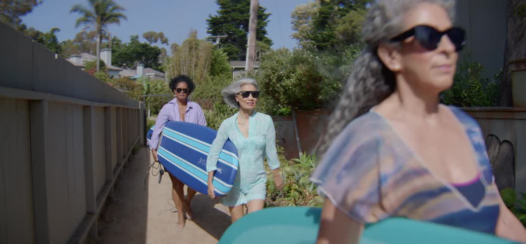A diverse group of active senior women friends happily walk down a sunny path with their surfboards to catch some waves