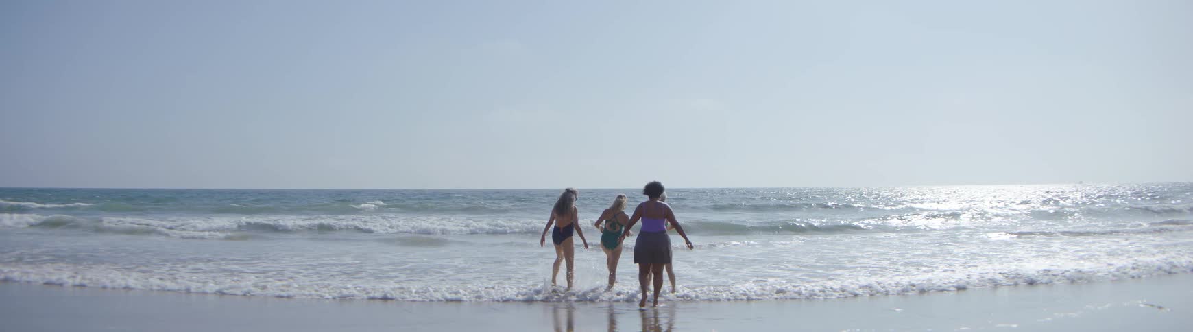 Slow motion shot of a group of senior women friends in swimsuits playfully walking into the ocean on a bright sunny day