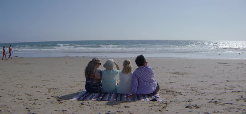 A diverse group of four women sit together on a blanket on the sand, peacefully watching the ocean waves on a sunny day