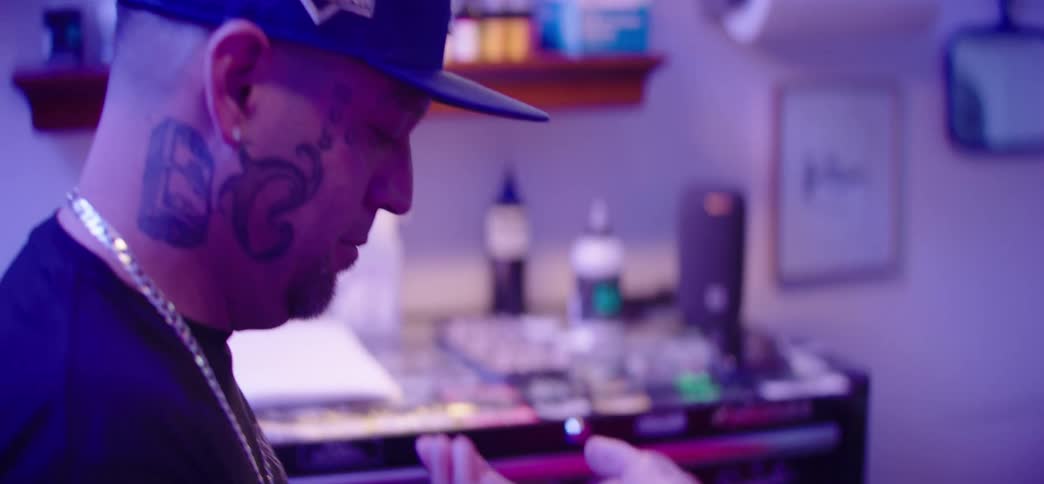 A tattoo artist with face tattoos prepares his tattoo machine before starting work on a client in a purple-lit studio