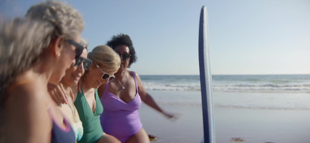 Cheerful senior women friends laugh and embrace while one practices balancing on a surfboard during a lesson at the beach