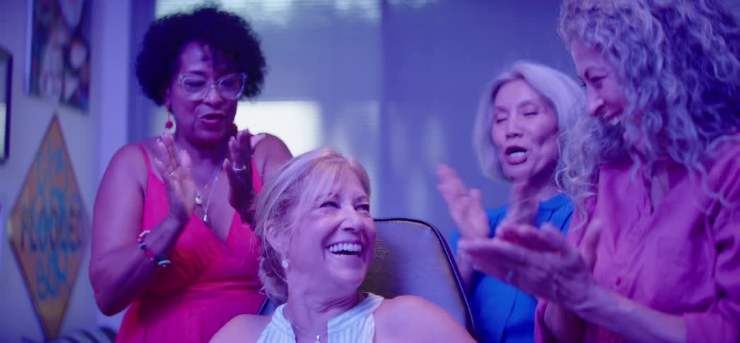 A group of diverse senior women friends celebrate a victory together, laughing, cheering, and clapping with excitement in a neon-lit room