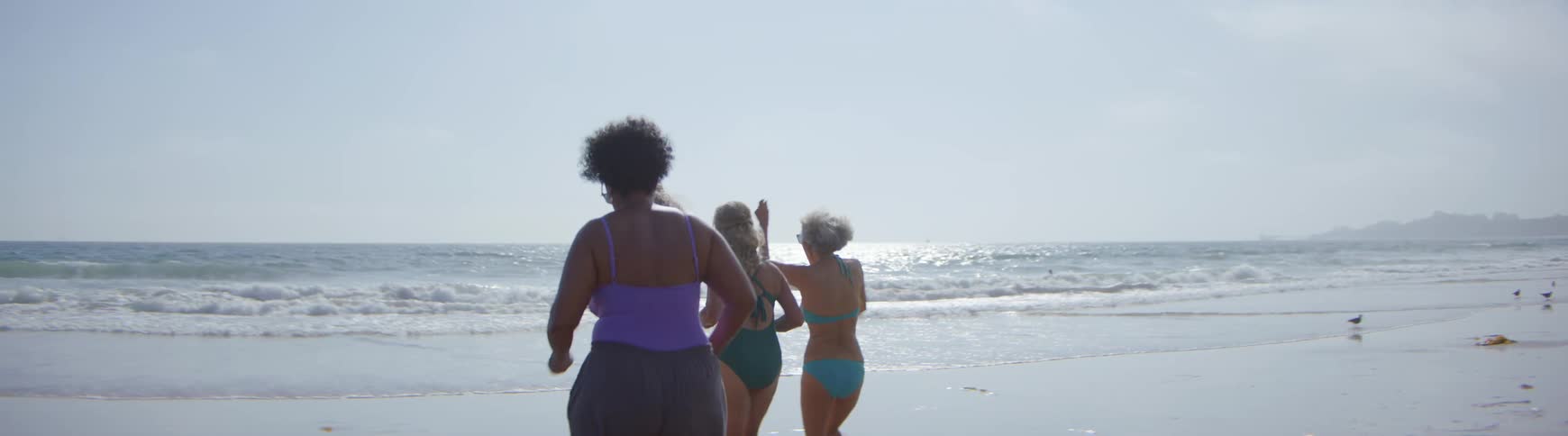 A group of diverse senior women in swimsuits happily run into the ocean together in slow motion on a sunny day