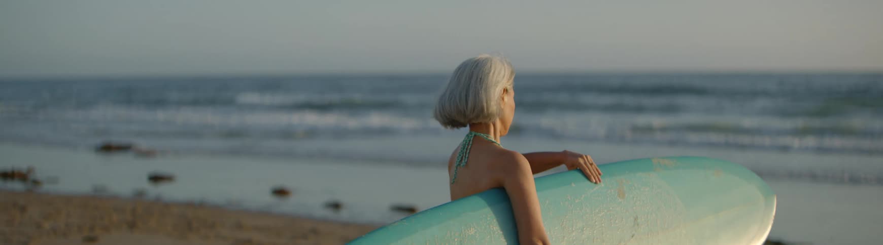 A graceful older woman in a bikini carries her surfboard in slow motion as she walks along the beach towards the gentle ocean waves