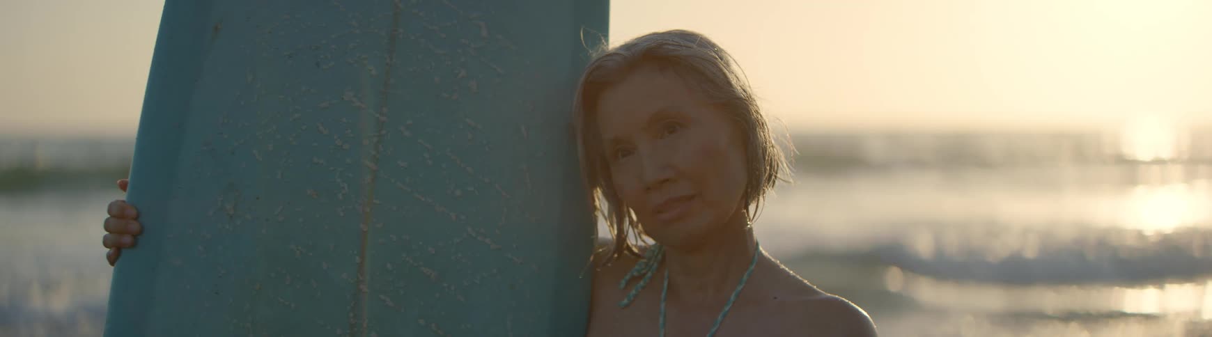 A serene senior Asian woman with wet hair rests her head against her surfboard, peacefully enjoying the golden light of sunset at the beach