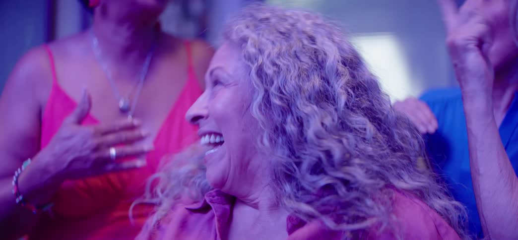 A radiant mature woman with curly gray hair laughs and dances with her friends at a party under vibrant purple lights