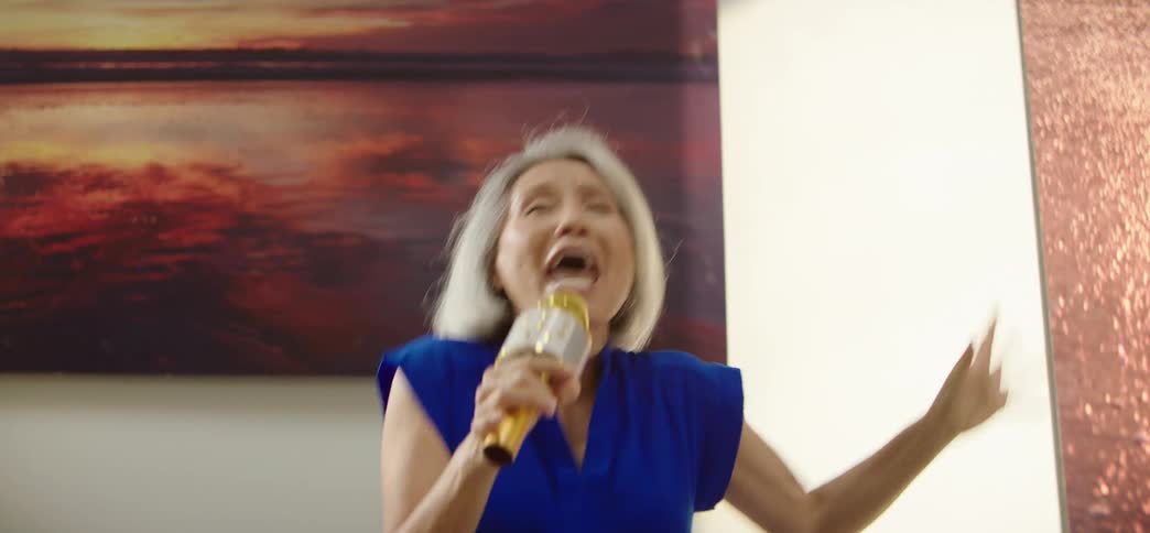 An energetic senior woman in a blue dress enthusiastically jumps on the couch and sings karaoke while her friends cheer her on in slow motion
