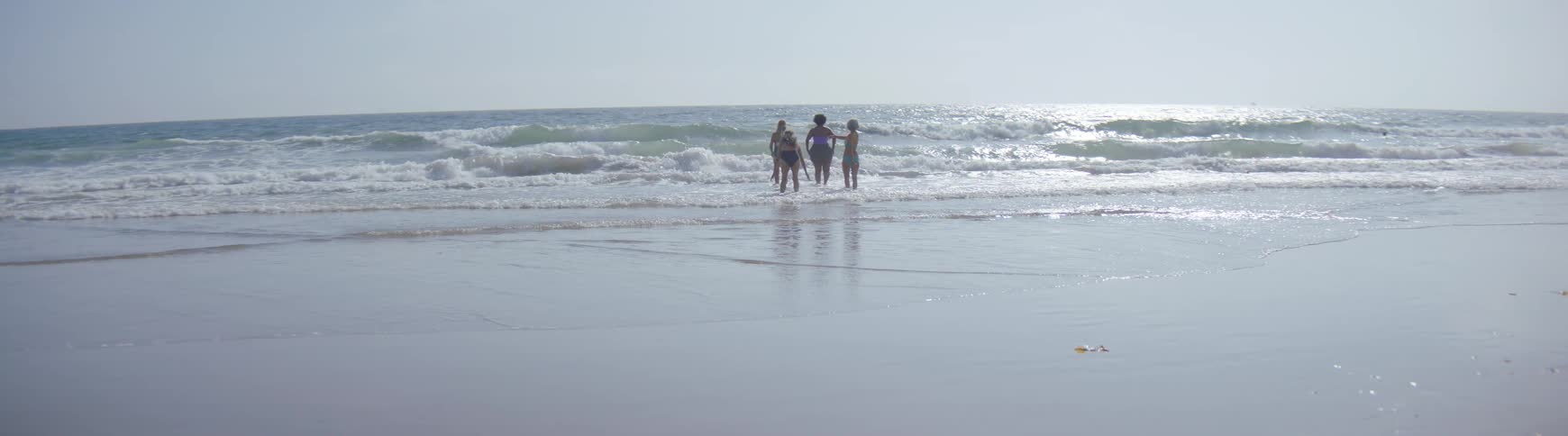 A group of four women in swimsuits enjoy a sunny day at the beach, walking into the ocean waves in slow motion