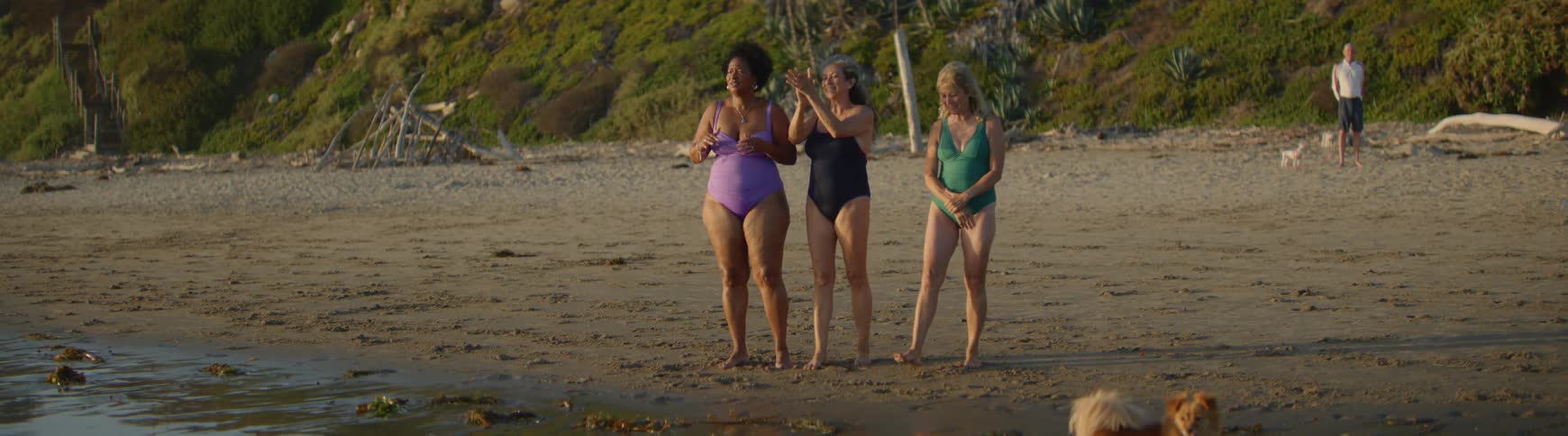 Three happy and diverse senior women friends in one-piece swimsuits stand on the beach and cheer for someone in the water