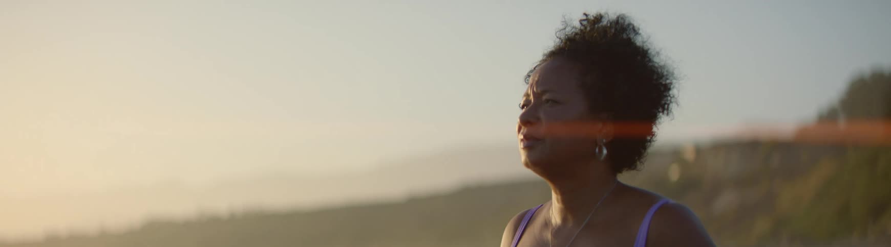 A mature Black woman stands in quiet contemplation as the sun sets, with a soft lens flare illuminating her profile