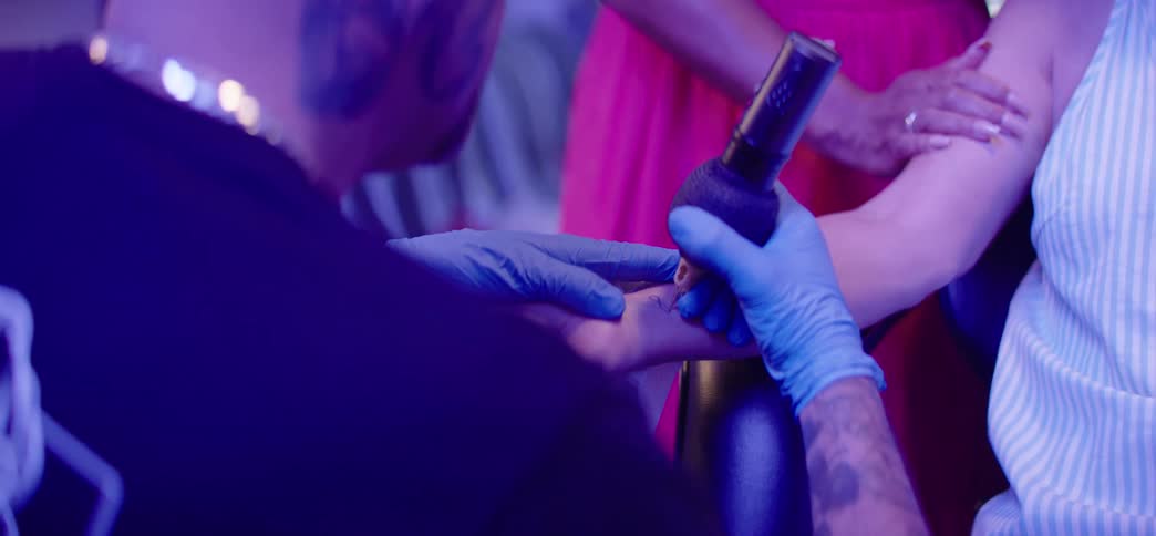 A joyful senior woman gets a tattoo on her wrist from a professional artist as her supportive friends cheer her on