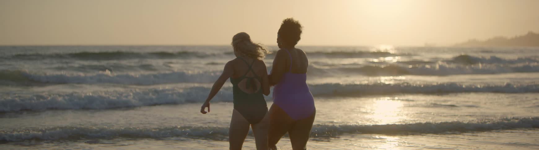 Two diverse female friends run into the ocean at sunset in slow motion