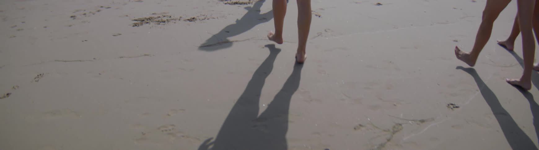 In slow motion, the bare feet and long shadows of several people glide across the sand as they run along the beach