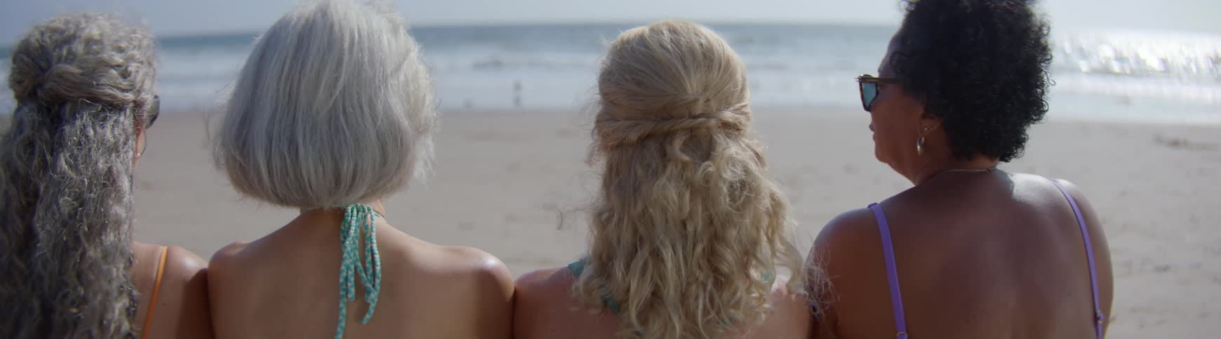 A back view of three diverse senior female friends in swimsuits relaxing on the beach and looking at the waves