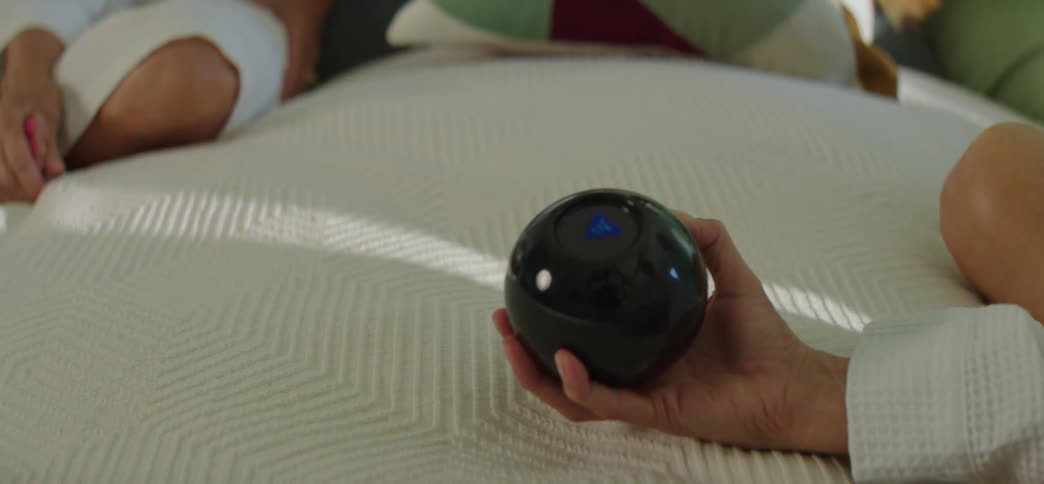 Close up of a hand shaking a Magic 8-Ball on a white bedspread, waiting for the fortune-telling toy to reveal an answer