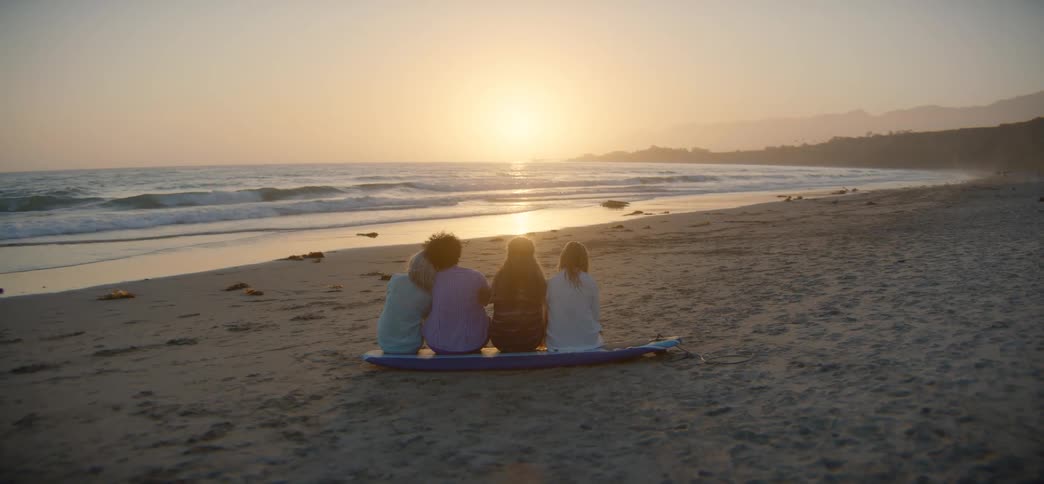 Rear view of four friends sitting together on a surfboard on the sand, peacefully watching the sun set over the ocean waves