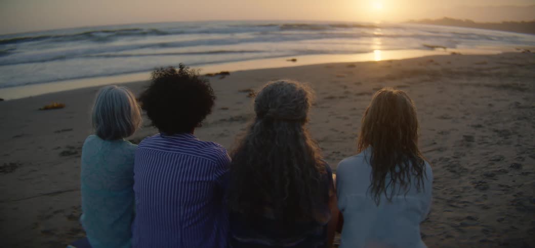 Four diverse female friends sit together on a sandy beach, peacefully watching the golden sunset over the ocean