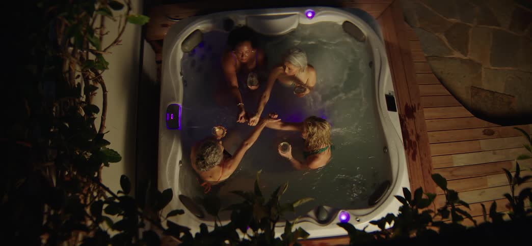 Overhead view of four diverse women holding hands and drinking wine while relaxing together in a bubbling hot tub at night