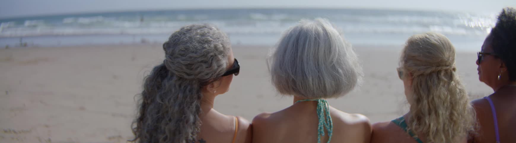 Back view of three diverse mature women relaxing on the beach, with one resting her head on her friend's shoulder in a moment of quiet companionship