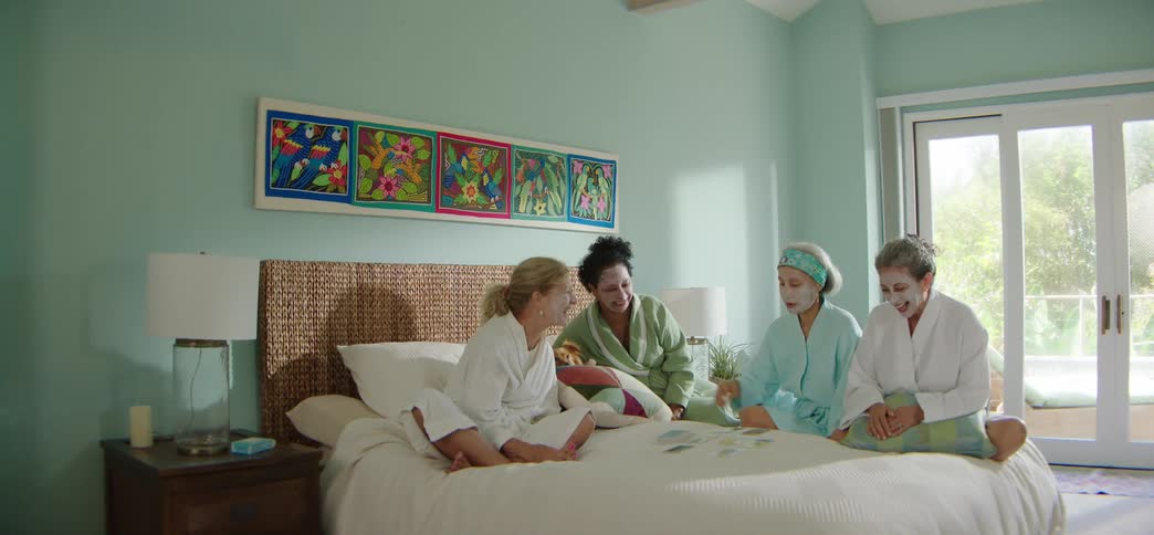 A diverse group of women in bathrobes and face masks laugh together while reading tarot cards on a bed during a spa day