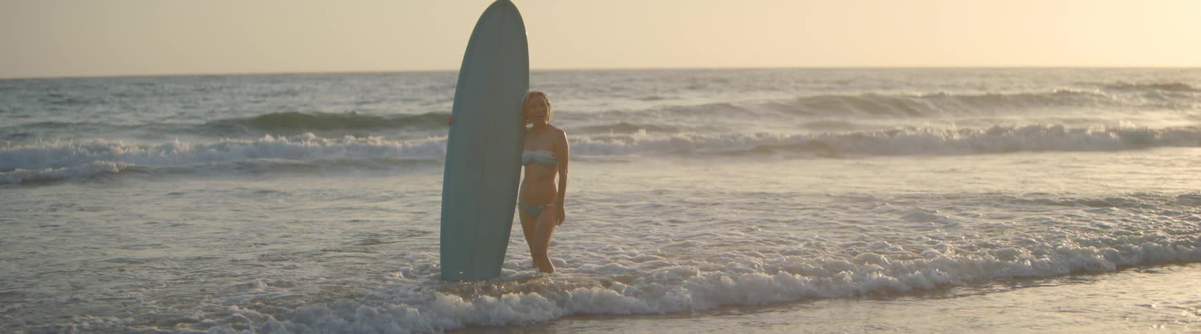 A smiling woman in a bikini holds her surfboard and poses in the gentle ocean waves at sunset