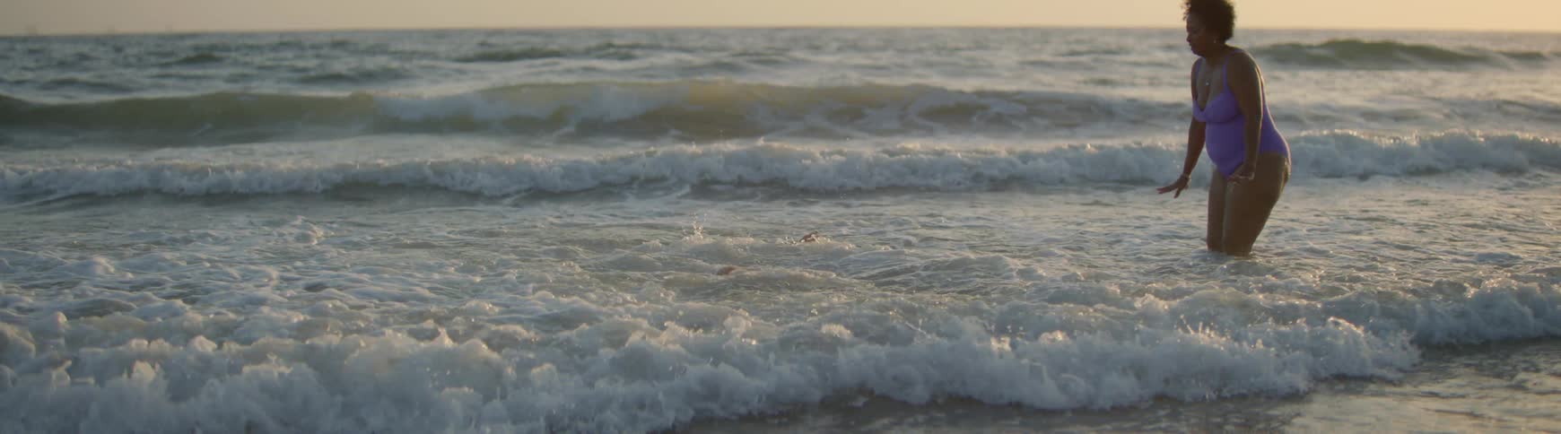 In slow motion, a woman playfully sits in the shallow ocean water, raising her arms just before a wave happily crashes over her