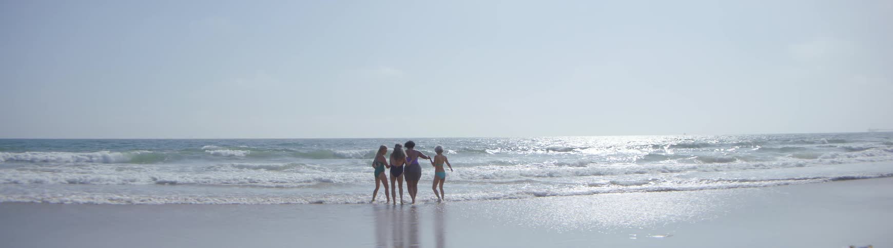 Diverse group of happy women hold hands and playfully walk into the ocean waves on a sunny beach