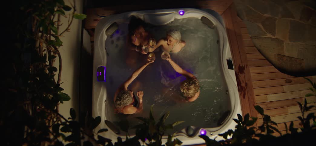 A high-angle view of four friends making a celebratory toast with wine while relaxing in a bubbling outdoor hot tub at night