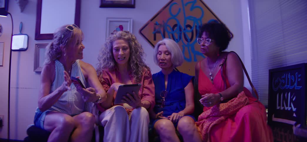 A fun group of diverse older women friends sit on a couch, laughing and talking while taking a selfie together with a smartphone