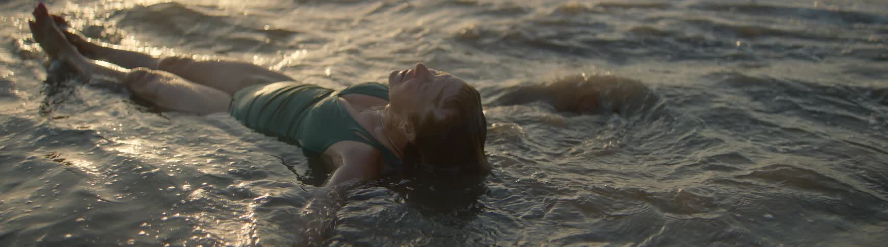 A serene woman in a swimsuit floats peacefully on her back in the gentle ocean waves before sitting up to enjoy the golden sunset