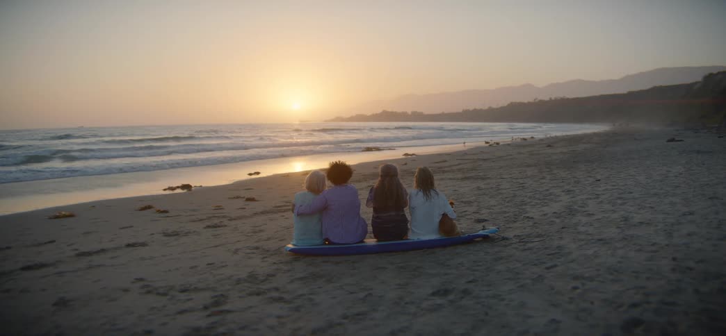 A group of four friends sit together on a surfboard on the beach, watching the beautiful sunset over the ocean