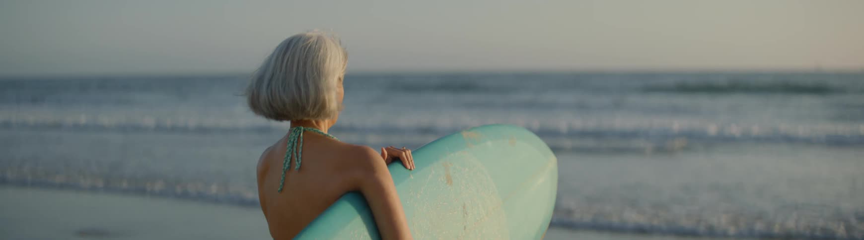 A senior woman with grey hair holds a surfboard while looking out at the ocean waves