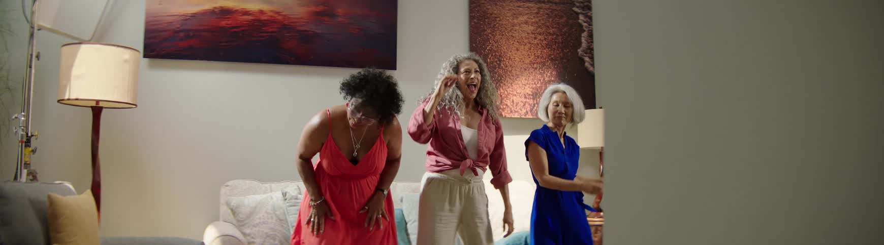A trio of diverse senior women laugh and dance with carefree energy in a bright living room, captured in slow motion