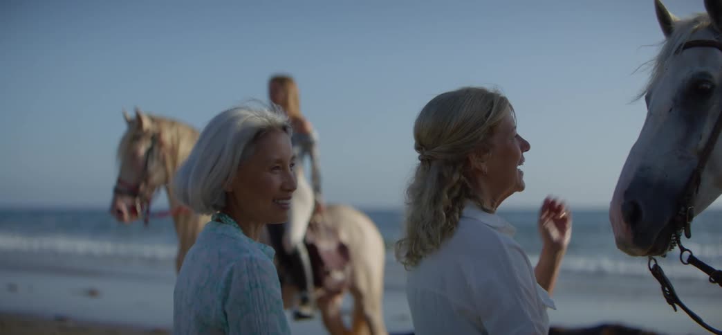 Smiling older women pet a beautiful white horse on the beach as the camera focuses on a young rider looking at the ocean