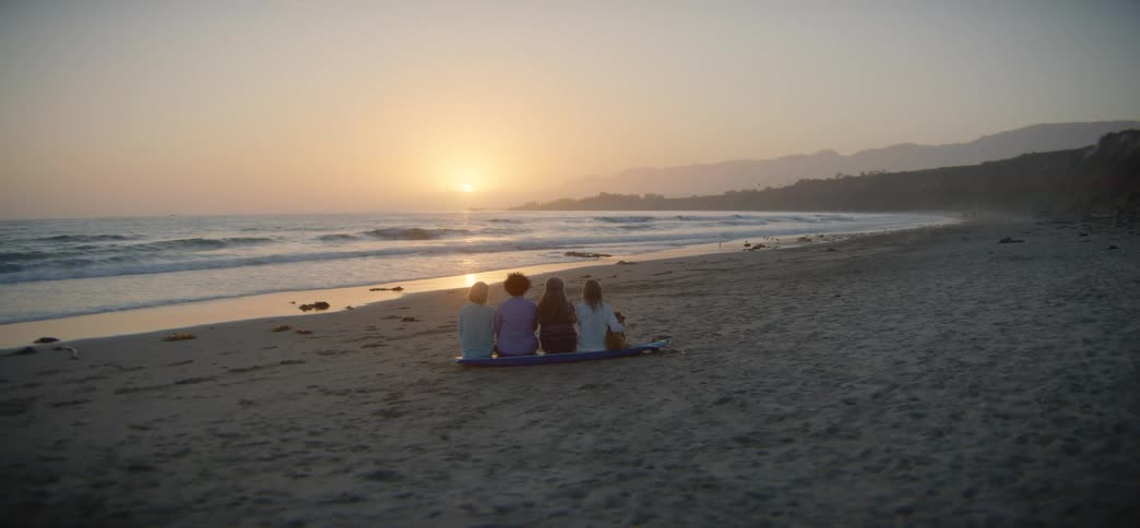 A slow focus pull reveals four friends sitting on a surfboard, peacefully watching the golden sunset over the ocean