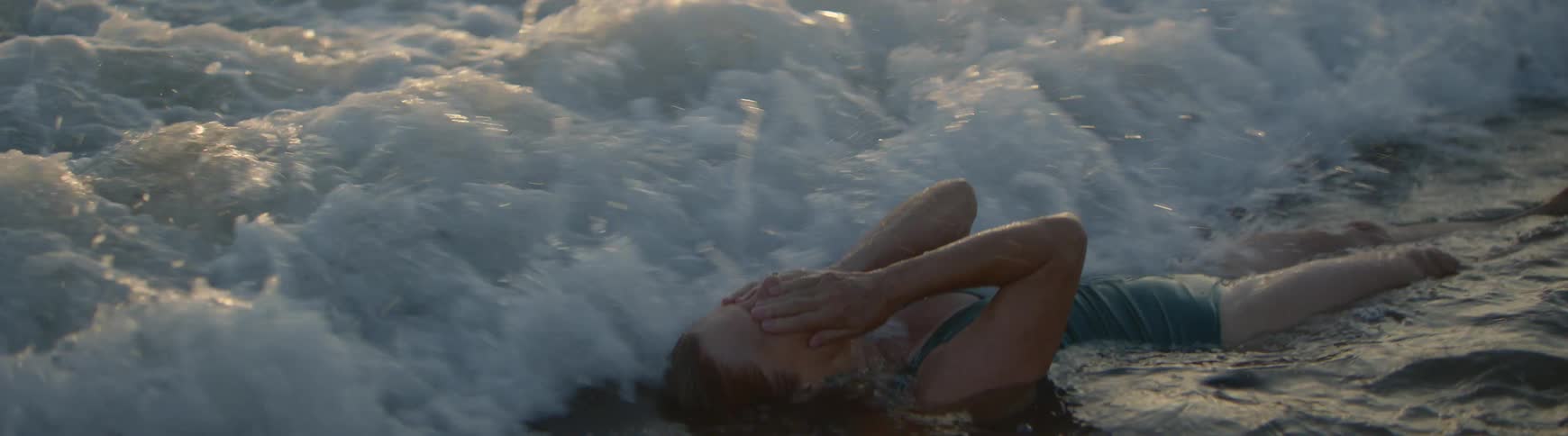 A woman joyfully floats on her back in the shallow ocean water at sunset as a wave washes over her in slow motion