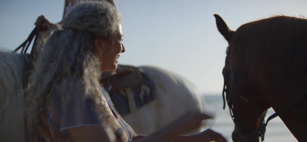 A smiling woman with long grey hair affectionately pets a brown horse on the face at the beach during a beautiful sunset