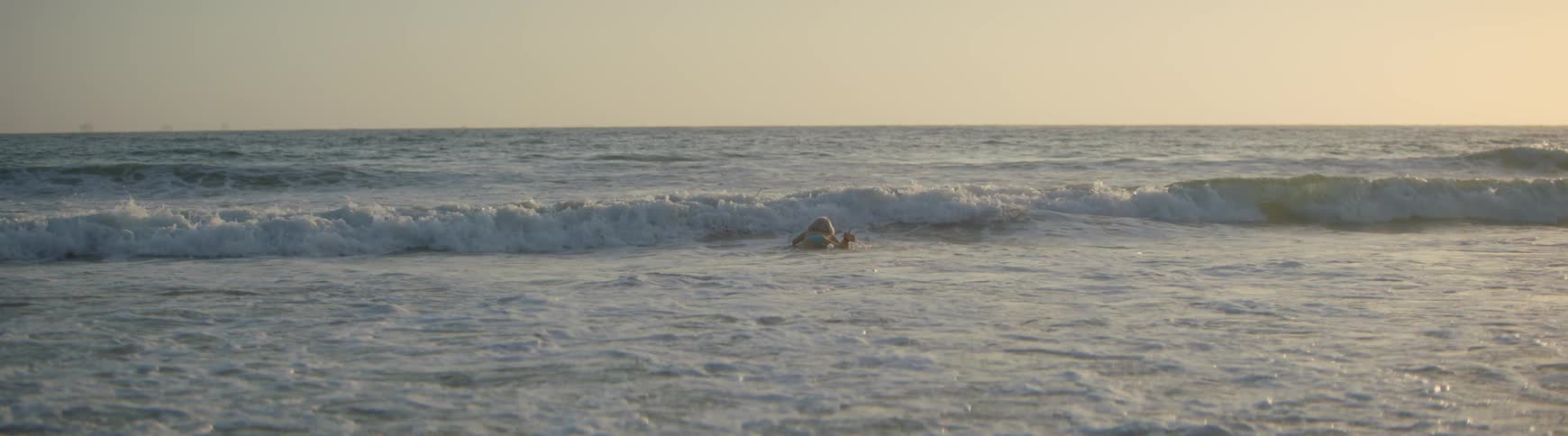 A female surfer carries her board into the ocean and paddles over the waves in slow motion during a beautiful sunset