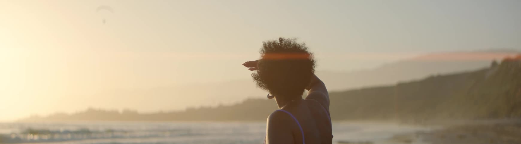 A contemplative woman in a swimsuit watches the ocean from the beach during a hazy golden hour, shielding her eyes to get a better look