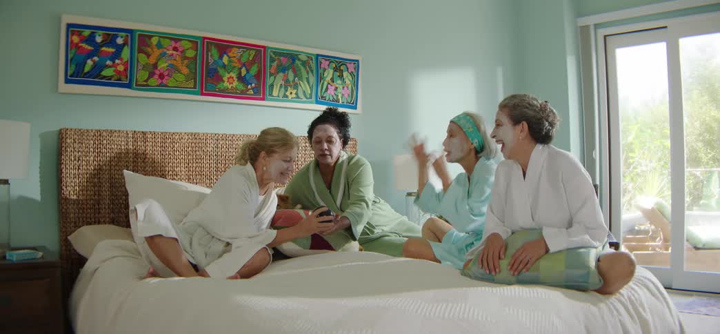 A diverse group of women in bathrobes and facial masks laugh together while relaxing on a bed during a spa day