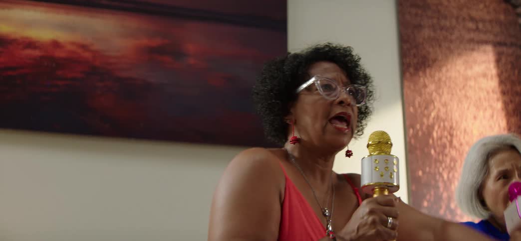 A senior woman passionately sings karaoke into a toy microphone in slow motion during a party at home with her friend